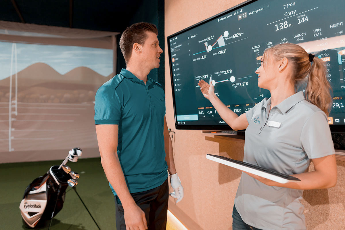 Golftrainerin erklärt einem Gast im Indoor-Golfsimulator des THE GRAND Ahrenshoop die Schwunganalyse am Bildschirm.